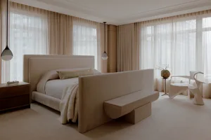 A minimalist bedroom featuring neutral-toned bedding and drapery in front of large windows with sheer curtains.