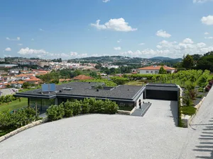 A modern villa with concrete exterior and a sunlit driveway overlooking a town.