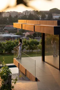 A woman stands on a modern veranda with glass panels and concrete flooring, overlooking a cityscape at sunset.