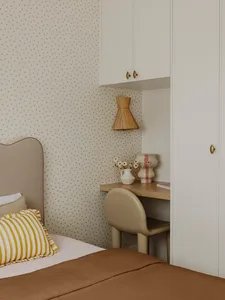 A minimalist bedroom with a bed featuring a neutral-toned fabric and a striped pillow, a desk with a vase of flowers and a la