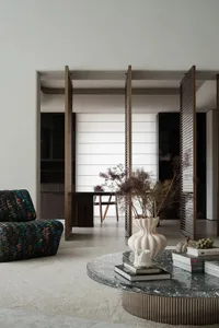 A modern minimalist living room with a round marble coffee table, a colorful patterned chair, and a white vase with dried pla