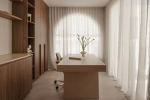 A modern minimalist office space featuring a wooden desk, white fabric curtains, and a vase with green plants.