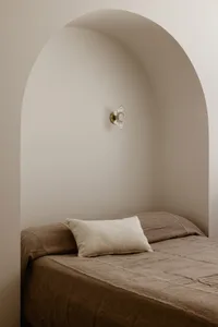 A minimalist modern bedroom with a bed covered in neutral textiles, framed by an archway and a sconce fixture.