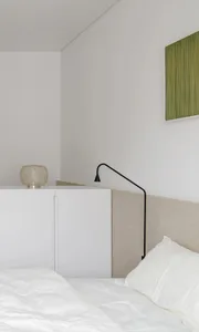 A minimalist white bedroom with a bed, nightstand, and green artwork on the wall.