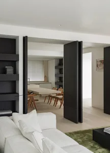 A modern living room with concrete floor, wooden furniture, and white cushions on a green rug.