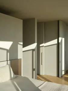 A modern minimalist bedroom with white walls and furniture, featuring sunlight shadows.