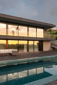Modern residential architecture featuring glass walls and wooden deck with a swimming pool and sunset view.