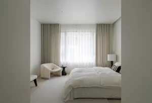 A minimalist bedroom featuring neutral-colored furniture and curtains, large windows with white sheer curtains, and a white b