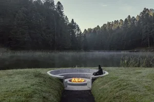 Concrete firepit surrounded by grass with a person sitting next to it in a serene forested landscape.