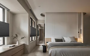 A modern minimalist bedroom featuring neutral-toned walls, a white bed with dark gray accents, and a natural light-filled win