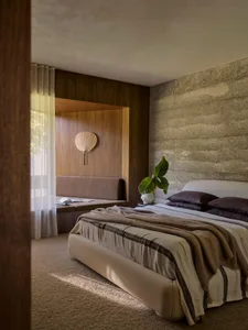 A minimalist bedroom featuring concrete and wood materials, a white bed with a striped blanket, and a wooden bench against th