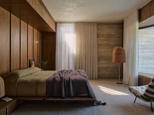 A modern bedroom featuring wooden walls and a concrete accent wall, with a bed and chair, and sunlight streaming through the