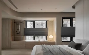 A modern minimalist bedroom with light wood flooring, a white bed with grey throw blankets, and a wooden cabinet with open sh