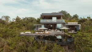 Modern vacation home with glass and concrete exterior, surrounded by lush greenery.