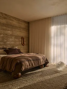 A minimalist bedroom with a concrete wall, a large window with sheer curtains, and a bed with a brown blanket and pillows.