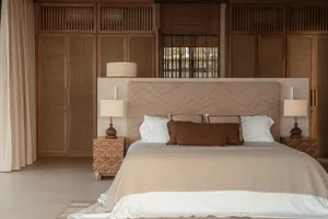 A minimalist bedroom featuring a taupe upholstered bed with matching comforter, surrounded by wooden furniture and a large wi