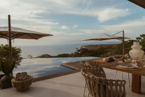Outdoor dining area with wooden furniture, glass infinity pool, and fabric parasols overlooking the ocean.