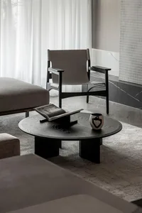 A minimalist living room scene with a round black coffee table, an open book, and a ceramic vase. A grey upholstered armchair
