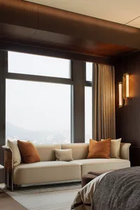 A modern bedroom featuring a beige leather couch with orange and beige throw pillows, large windows with views of a cityscape