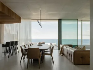 A modern living room with concrete and glass walls, large windows overlooking the ocean, and minimalist furniture.