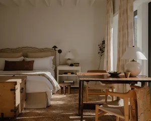A minimalist bedroom featuring wooden furniture and neutral tones with a sunlit area and a large window.