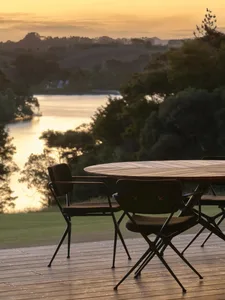 Modern outdoor seating area with chairs and table on a timber deck overlooking a serene lake at sunset.
