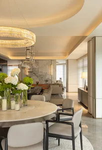 A modern residential dining room featuring a round marble dining table, white leather chairs, and a crystal chandelier.