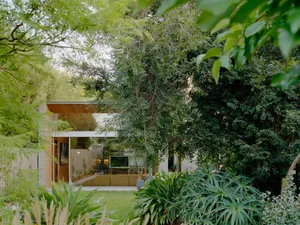 A modern residential exterior featuring wooden panels, glass walls, and concrete floors surrounded by lush greenery.