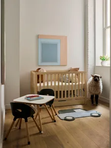 Minimalist children's bedroom featuring a crib, small table, chairs, and a woolly sheep against a window with a plant.