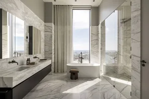 A modern bathroom featuring a freestanding bathtub, white marble flooring and walls, and a large window overlooking a citysca
