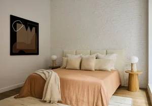 A minimalist bedroom featuring a bed with beige bedding and a wooden headboard, with a modern abstract painting on the wall a