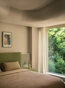 A minimalist bedroom featuring a bed with neutral fabrics, a green headboard, and a window with curtains overlooking greenery