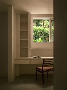 A white study room with a built-in desk, shelves, an open book on the desk, and a mid-century chair.