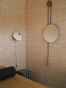 A minimalist bedroom featuring plywood walls, a bed with brown bedding, and a wall-mounted lamp and mirror.