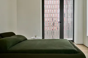 Modern minimalist bedroom featuring a green upholstered bed with a transparent side table and view of a cityscape through lar