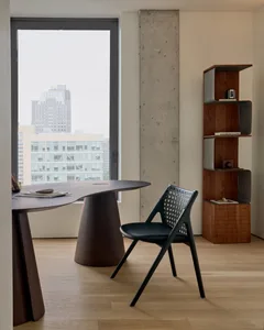 A minimalist office space featuring a modern, curved table with wooden legs and a black chair with a woven backrest. The room