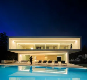 A modern, sleek house with a glass wall and concrete patio features a serene outdoor area with a clear blue sky and lush gree