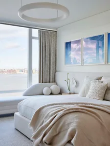 A minimalist residential interior featuring a white fabric bed with gray curtains and framed artwork on the wall.