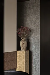A wooden vase with dried flowers placed on a wooden standing base against a textured grey wall.