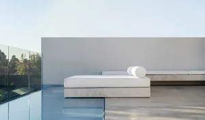 Modern, minimalist concrete seating area by a pool with white cushions and a cylindrical pillow.