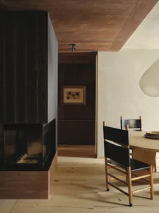 A modern residential dining room featuring a wooden floor, black chairs, and a large window with a view of the natural surrou