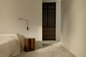 Minimalist bedroom with white walls, a bed with a cream blanket, a wooden stool, and a large closet with sliding doors.