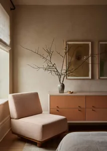 A minimalist bedroom featuring a pink armchair, a chest of drawers, and a bed with a white quilt, decorated with a vase of bl