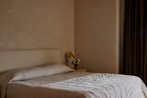 A minimalist bedroom with a bed, a side table, a lamp, and a vase with flowers, all covered in light-colored textile material