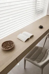 A modern minimalist home office features a wooden desk with an open book, bowl of peanuts, and a mug, alongside a chair and a