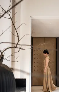 A woman in a beige dress stands in a minimalist home interior with a textured, neutral-toned wall and a dark, sculptural door