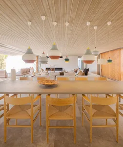 A modern living and dining room featuring a concrete ceiling and walls with wooden furniture and colorful pendant lights.