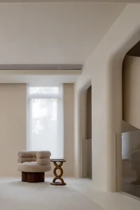 A minimalist living room with neutral-toned walls, a single plush armchair with pillows, a small wooden side table, and a lar