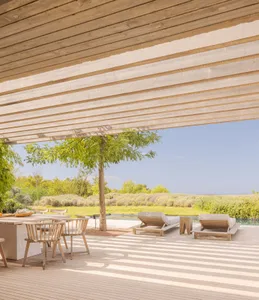 A modern outdoor living space featuring wooden deck and lounge chairs under a sheltered pergola overlooking greenery.