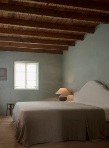 A minimalist bedroom with a bed covered in fabric, a wooden headboard, and a nightstand lamp.